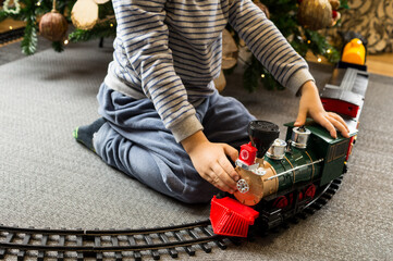 A boy sits cross-legged on a gray carpet, looking intently as a toy train travels along tracks encircling a beautifully decorated Christmas tree. © Phoscar