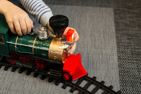 A boy kneeling on a gray carpet, wearing a striped long-sleeve shirt, focuses on assembling a toy train set with tracks in front of a decorated Christmas tree.