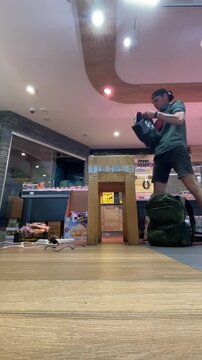 man standing beside wooden stool inside small retail shop retrieving item from dark backpack, low floor-level viewpoint emphasizes wood-look flooring, centered shop