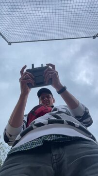 adult holds remote controller overhead with both hands, thumbs near control sticks while looking upward beneath visible mesh safety net in soft overcast outdoor
