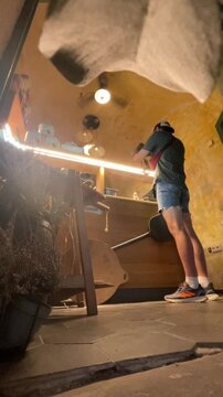 young adult customer standing at wooden counter in rustic cafe, holding handheld device toward countertop while inspecting or photographing items, scene shot from