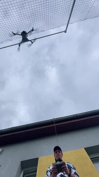 drone hovers above pilot near building facade under overcast sky, with blurred rotors and operator holding remote controller at ground level below in soft daylight
