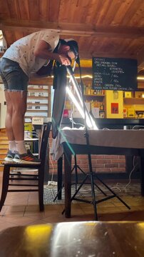 person balances on chair while aiming camera toward illuminated tabletop, using bright led tubes and cozy warm interior lighting for focused product setup indoors