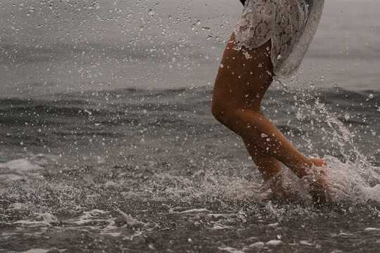 Person walks in shallow water while playing and splashing during the sunset. A person splashes in the shallow water at the beach. Waves hit their legs as the sun sets.