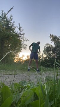 young man performs upward arm stretch on dirt path at dusk, hands clasped overhead and body slightly leaning back in energetic or victorious pose