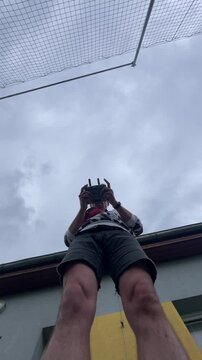 person stands on rooftop edge, holding handheld remote controller with both hands, seen from low angle under cloudy sky, with soft daylight and muted urban tones