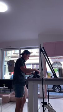 man leans and crouches at tall worktable to photograph items on table, holding camera to eye and adjusting controls with intent focus, with man, photographing man