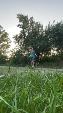male athlete performing jump rope exercise beside quiet roadside at dusk, captured from very low angle that emphasizes tall foreground grass and deep depth, with