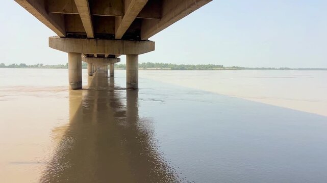 A close-up view of a vast river filled with flowing water and natural surface eddies, as it flows calmly beneath a sturdy bridge. 4k video