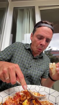 close-up motion of adult man eating at window table, lifting fork with saucy cheesy skillet topping while holding partially eaten sandwich in other hand and, with