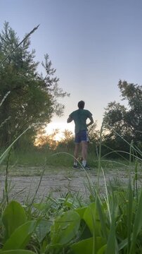 single young male in green t-shirt and blue athletic shorts performs upward stretch, steps forward, then pauses with hands on hips on dirt path at sunset, low male