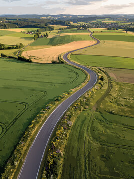 A scenic winding country road passes through expansive green and golden agricultural fields under a cloudy rural sky.