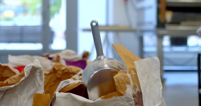 metal scoop with vertical handle protrudes from torn brown and white paper bags on counter of bakery or commercial kitchen, foreground cluttered with crumpled scoop