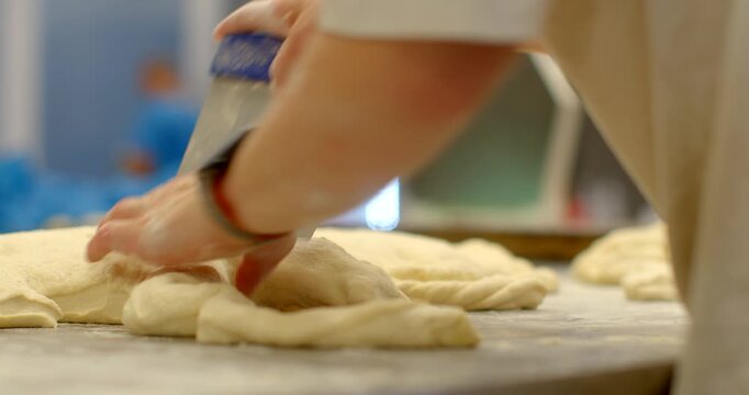 close-up view of baker hands pressing and portioning pale elastic dough, then arranging pieces on floured stainless worktop inside bakery kitchen, dusting flour