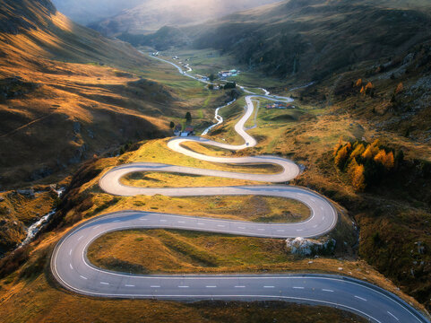 Aerial view of winding mountain road, river in alpine valley at sunset in autumn. Top drone view of road in Swiss mountain pass, yellow grass, trees in fall. Colorful landscape. Alps in Switzerland