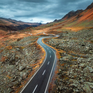 Aerial view of winding mountain road in the Swiss Alps, Switzerland. Rocky alpine landscape with dramatic cloudy sky and high mountains in fall. Top drone view of road in autumn. Transportation