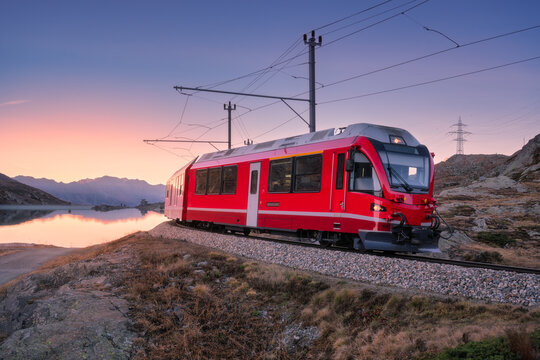 Bernina Express train near Lake Bianco in Switzerland at sunset, purple sky, clear reflections, mountain silhouettes and alpine terrain in evening light in the Swiss Alps. Red train at dusk. Transport