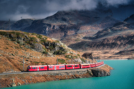 Aerial view of red Bernina Express train curving along the turquoise lake Bianco in the Swiss Alps, rocky hills and dramatic cloudy sky in autumn alpine mountain landscape. Top view of train in fall