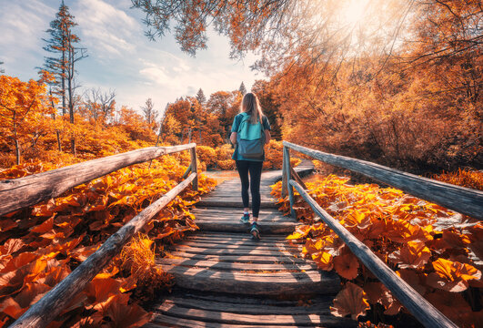 Young woman with backpack walking along a wooden path near stunning waterfalls, orange trees in Plitvice Lakes national park, Croatia at sunset in autumn. Sporty girl in forest in fall. Hiking, travel