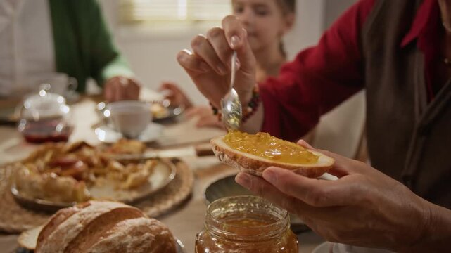 Senior Caucasian woman spreading fruit jam onto bread slice at breakfast table with family. Footage suitable for organic food advertising, bakery marketing, or morning lifestyle campaigns