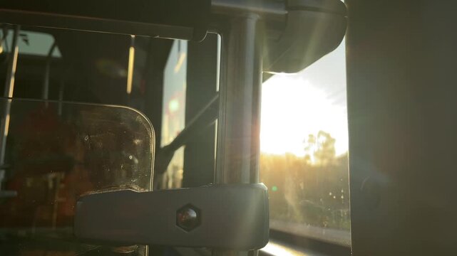 tight eye-level closeup of polished vertical support pole with molded plastic clamp and scratched transparent divider panel inside bus, sun streaming through side