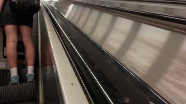 low-angle close-up showing glossy black escalator handrail in sharp foreground and passenger lower legs standing on ribbed steps in left background, reflective rail