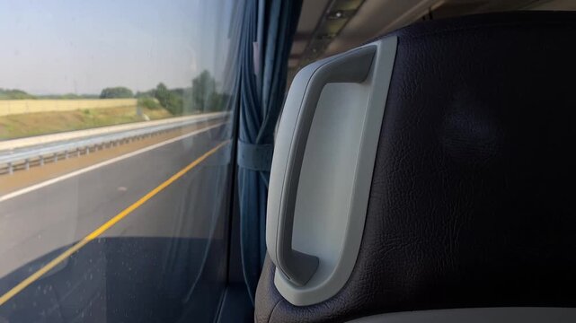 close-up view of plastic grab handle mounted on dark textured seatback inside coach with tied blue curtain beside window, multilane highway and metal guardrail bus