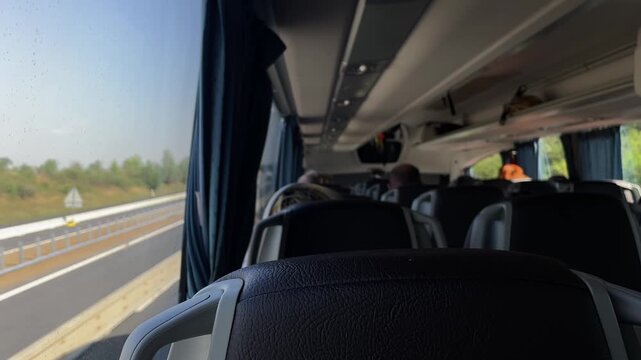 forward-facing interior shot from passenger eye level showing top of leather seatback in foreground, rows of reclining coach seats and overhead luggage racks, with