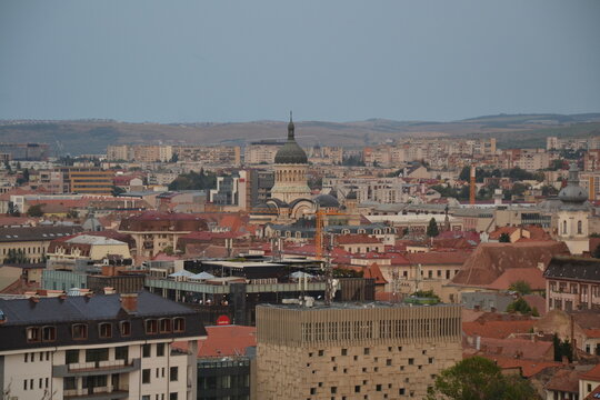 Panoramic view of Cluj-Napoca, Romania, with red-tiled rooftops, historic church towers and traditional architecture, set against rolling hills and the Carpathian Mountains under a cloudy sky.
