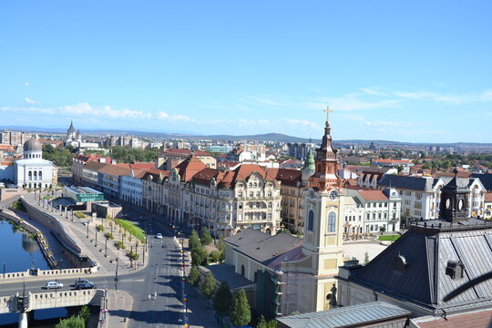 Aerial view of Oradea, Romania, featuring the historic city center, colorful rooftops, elegant architecture, Crișul Repede River, and distant Apuseni Mountains under a clear sky.