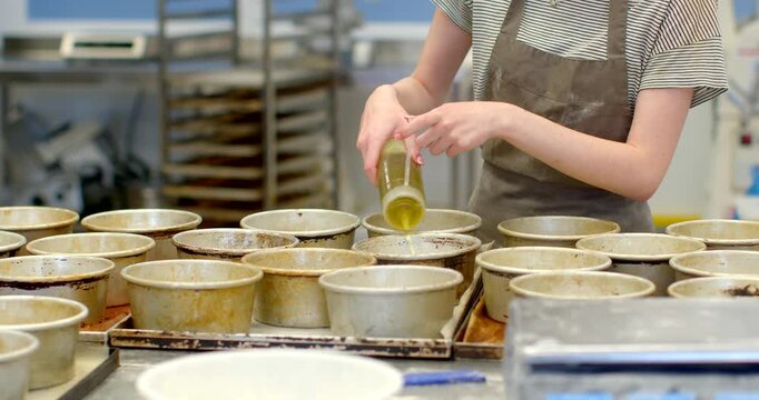close-up of bakery worker hands pouring oil from squeeze bottle into rows of round metal baking molds on stainless prep table, worker visible from torso wearing