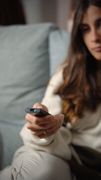 Close-up of a young Latina woman&rsquo;s hand using a television remote while sitting on a sofa, highlighting home entertainment, media choice, technology, and everyday leisure.