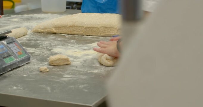 close-up view of bakery worker hands portioning and shaping dough on flour-dusted stainless steel worktable with larger loaf visible in background, digital scale
