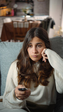 Young Hispanic woman sitting bored on her sofa while watching television, portraying downtime, home entertainment, media consumption, and an unengaged leisure moment.
