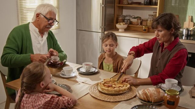 Senior Caucasian grandparents pouring tea and cutting homemade pie with granddaughters at kitchen table. Video ideal for intergenerational family bonding and retirement lifestyle campaigns