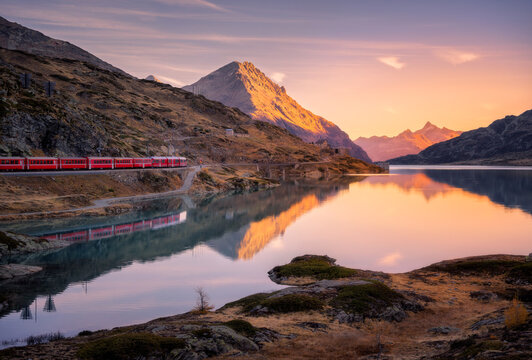 Aerial view of Bernina Express train near Bianco lake in the Swiss Alps at sunset, with glowing mountain peaks, water with reflections, rocky shore. Peaceful autumn landscape. Mountain train in fall