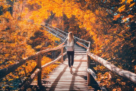 Young woman with backpack walking along a wooden path near stunning waterfalls, orange trees in Plitvice Lakes national park, Croatia at sunset in autumn. Sporty girl in forest in fall. Hiking, travel