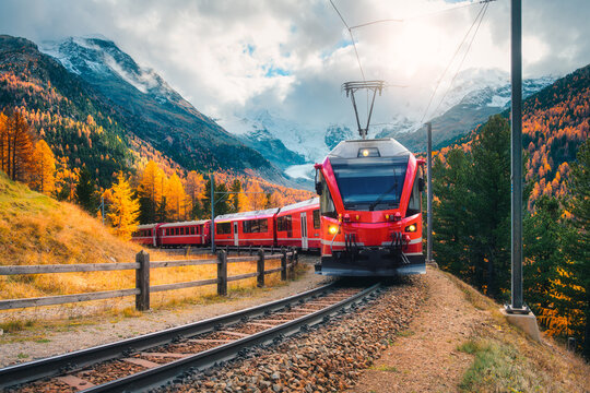 Red modern electric train traveling along a railway, winding through a stunning high mountains with snow-capped peaks and vibrant autumn trees in switzerland. Bernina Express in fall. Railway tourism