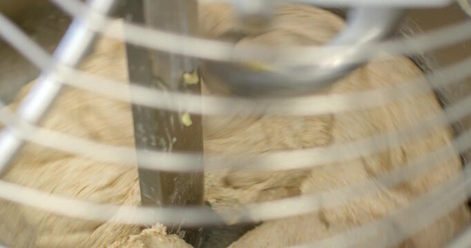 hydrated wholegrain dough being folded and consolidated around vertical stainless steel mixing blade inside commercial mixer bowl, seen through curved protective