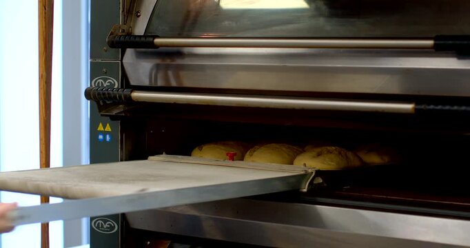 two bakers wearing pale work shirts place canvas-lined metal tray with rounded proofed dough into stainless steel commercial oven, hands gripping tray handle while