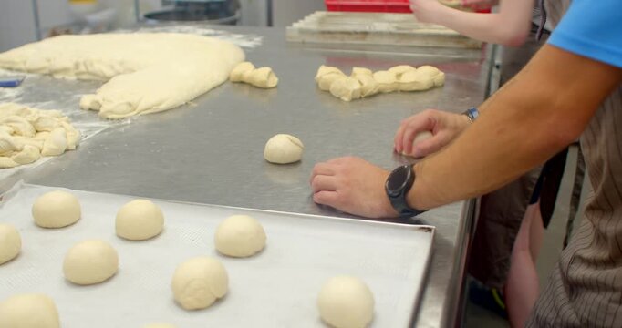 close-up view of bakers preparing bread dough on stainless steel counter, showing transformation from large bulk mass to evenly portioned round rolls, hands and