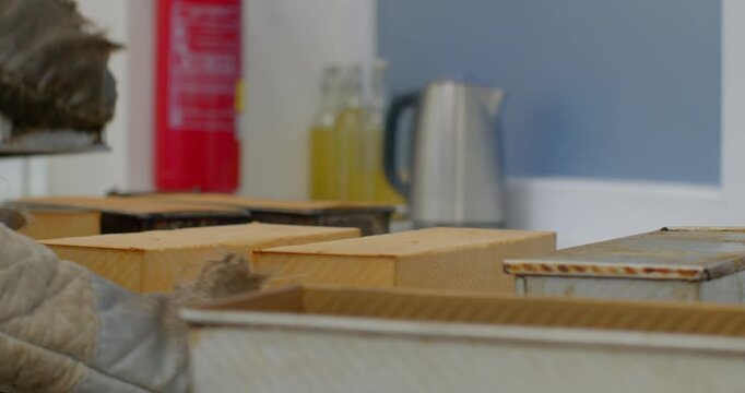 gloved hands of bakery worker place and adjust metal loaf pans beside rectangular bread loaves on countertop, quilted oven mitts, worn enamel tins, and wooden bakery
