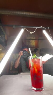 person preparing a drink, in stage with lighting and backdrop, with glass of red liquid, straw, microphone, from a front view perspective, with red, black accents.