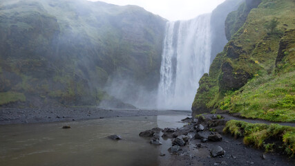 Obraz premium Majestic Skogafoss Waterfall plunges into a river surrounded by lush green cliffs in South Iceland, a famous filming location featured in films and television productions.