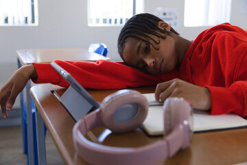 African male youth leaning on right arm at class desk, watching tablet, pen and pink headphones