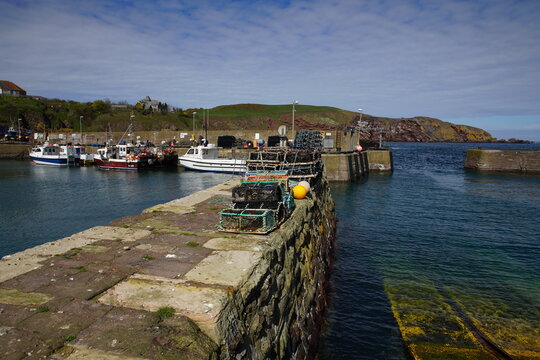 Fishing boats moored in St Abbs Harbour with stacks of lobster creels on the stone pier, Berwickshire, Scotland