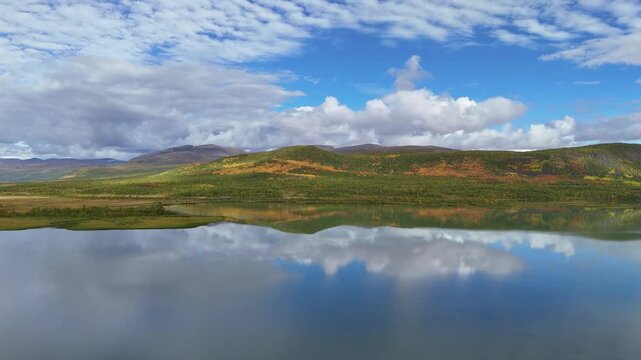 Aerial view of a pristine lake reflecting a dramatic blue sky and fluffy clouds in Arctic Sweden, surrounded by rolling hills with autumn foliage.