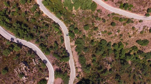 Bird's eye view of a serpentine two-lane road curving through dry Mediterranean hillside with scattered pine trees and rocky terrain, single car driving
