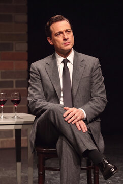  A man in a formal gray suit, white shirt, and dark tie sits on a chair. He holds himself calmly and confidently, one leg crossed over the other, his hands folded relaxedly over his knee.