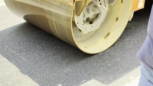 A yellow heavy road roller is seen working on a road construction and repair site, compacting a fresh layer of dark grey asphalt. The image showcases the construction equipment, steel drums, and the